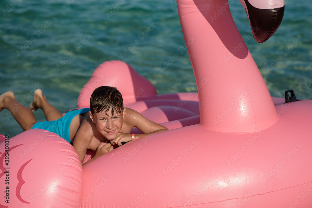 Cute European boy laying on huge inflatable pink flamingo in sea, he enjoying his vacations and smiling to camera.