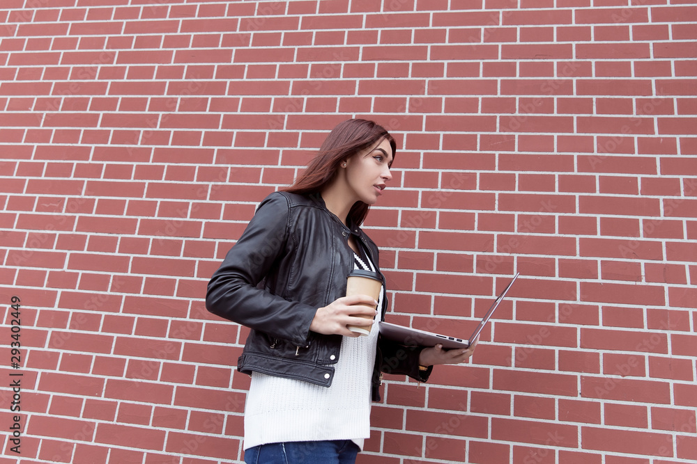 Red-haired girl in a black leather jacket works at the laptop against a red brick wall Red-haired girl in a black leather jacket works at the laptop against a red brick wall