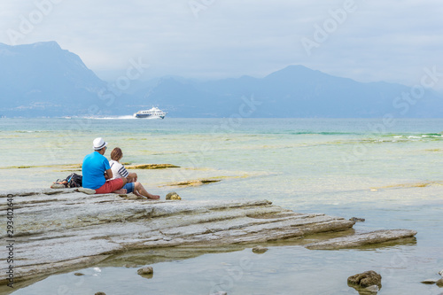 Zwei Urlauber sitzen am Strand und schauen auf ein Schiff