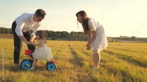 Dad and Mom teach daughter to ride bike. Mother and father play with small child on lawn. kid learns to ride bicycle. parents and little daughter walks in park. concept of happy family and childhood
