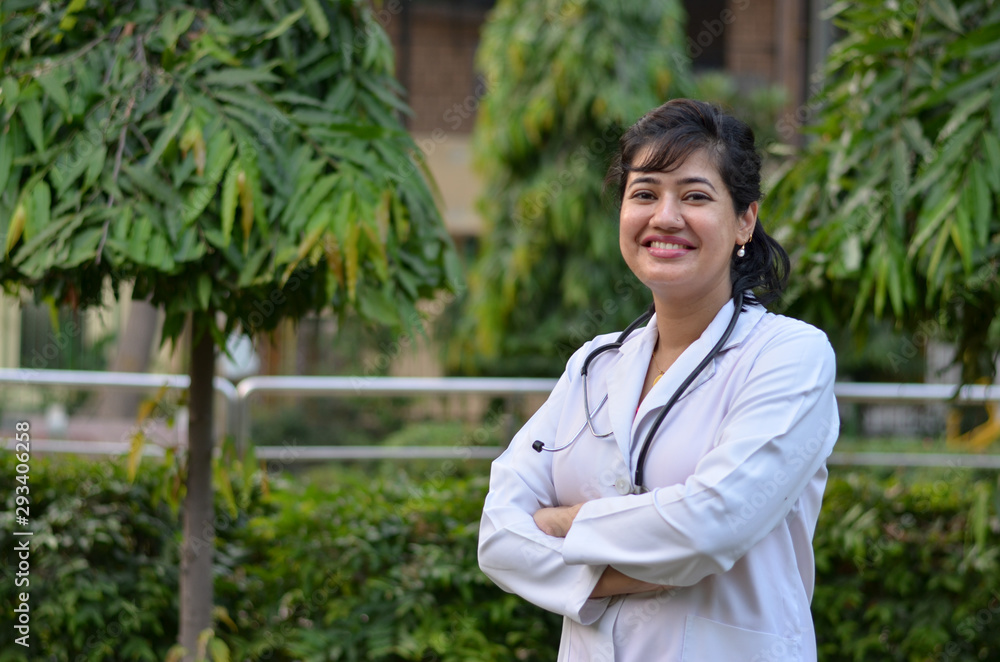 Portrait of a confident young female professional doctor wearing white coat and stethoscope in an outside setting in a park or hospital lawns in New Delhi, India Portrait of a confident young female professional doctor wearing white coat and stethoscope in an outside setting in a park or hospital lawns in New Delhi, India