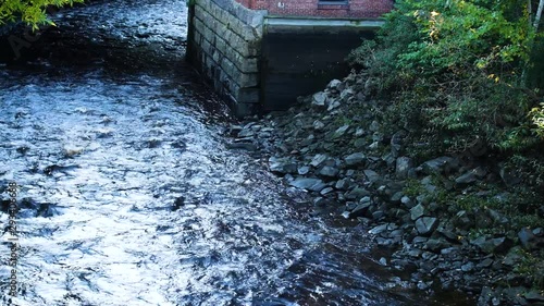 Video of Kenduskeag River water flowing past a building and riverbank with trees in the early morning light at Bangor, Maine. photo