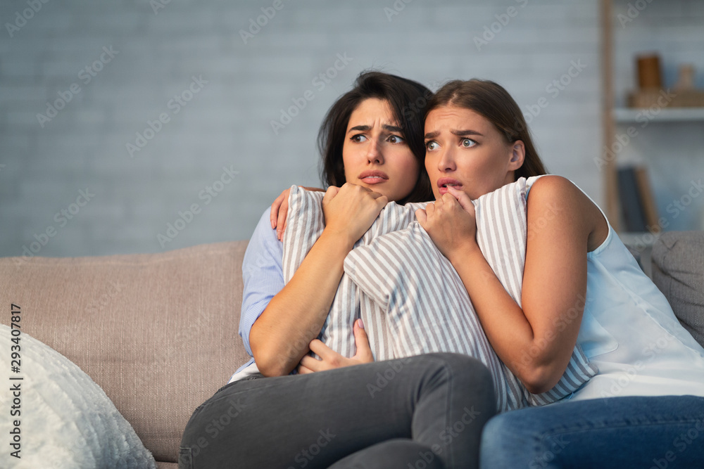 Two Scared Girls Hugging Pillows Sitting On Couch At Home Two Scared Girls Hugging Pillows Sitting On Couch At Home