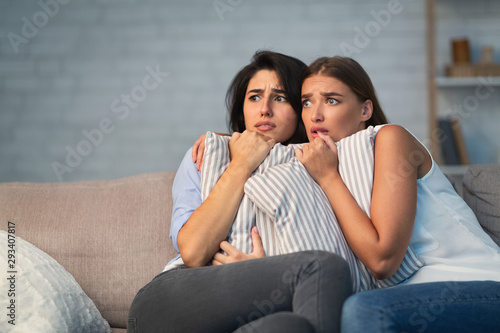 Two Scared Girls Hugging Pillows Sitting On Couch At Home