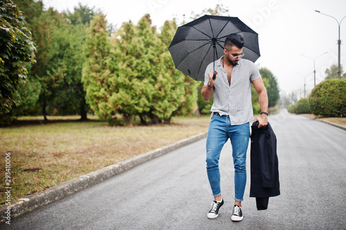 Fashionable tall arab beard man wear on shirt, jeans and sunglasses walking at park with umbrella and coat at hand.
