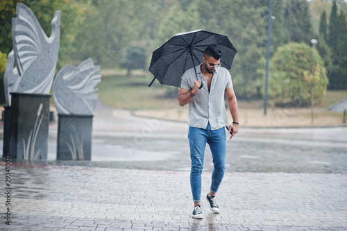 Fashionable tall arab beard man wear on shirt, jeans and sunglasses with umbrella posed at rain on park square.