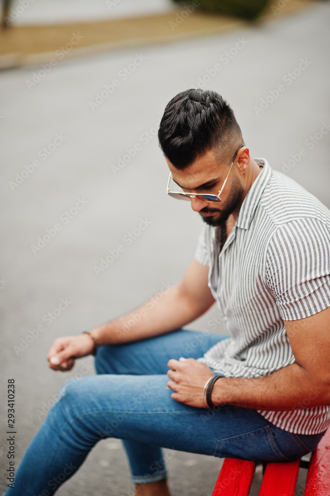 Fashionable tall arab beard man wear on shirt, jeans and sunglasses sitting on red bench at park. Fashionable tall arab beard man wear on shirt, jeans and sunglasses sitting on red bench at park.