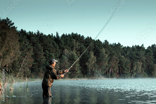 A male fisherman on the lake is standing in the water and fishing for a fishing rod. Fishing hobby vacation concept. Copy space. © Aliaksandr Marko