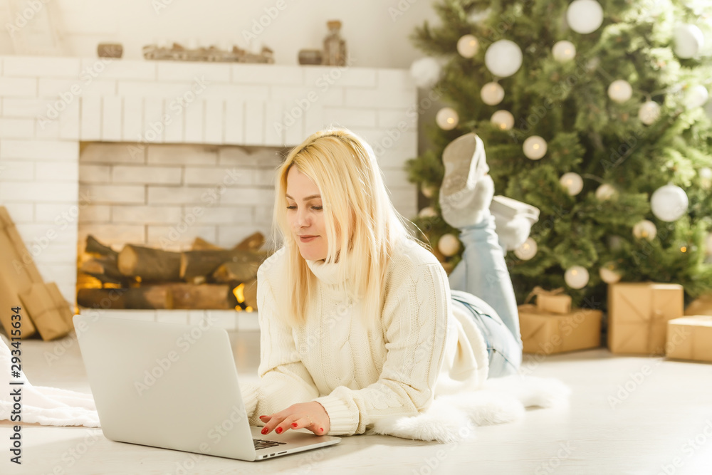 Young woman enjoying Christmas Eve sitting on the floor in front of a decorated Xmas tree in the living room using a laptop computer to maker online purchases.