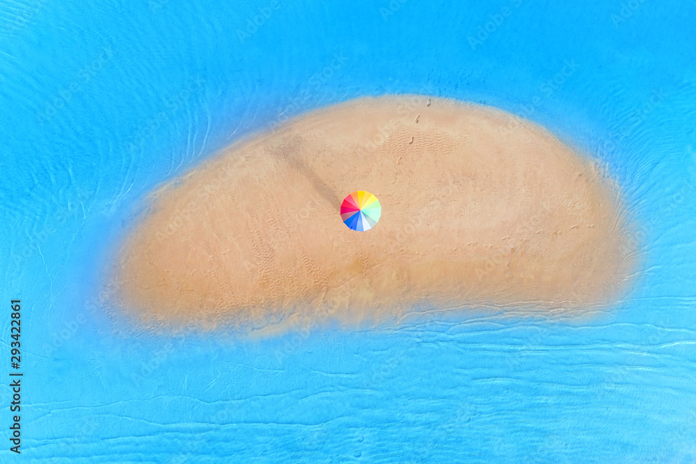 Small sandy island with someone under rainbow colored umbrella on it