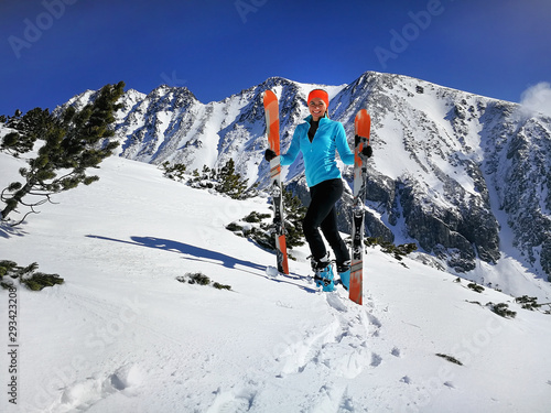 Young woman in light blue jacket standing, smiling happy holding with mountaineering ski and poles in hands, sun shines on snow, bright sky over mountains behind