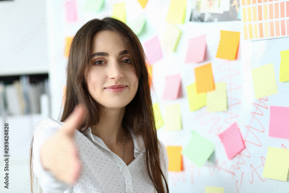 Portrait of gorgeous smiling creator standing near white board with colorful stickers and reaching tender arm to shake hands with friendly partner. Art design concept. Blurred background Portrait of gorgeous smiling creator standing near white board with colorful stickers and reaching tender arm to shake hands with friendly partner. Art design concept. Blurred background