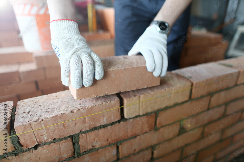 Close-up of prudent man wearing special protective gloves to prevent serious work injuries. Male hands putting brick on wall on big masonry. Building concept