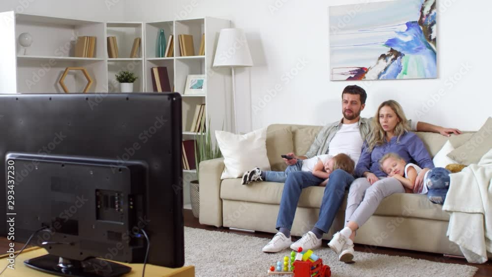 Tracking shot of couple sitting on sofa and watching TV as their little son and daughter sleeping on their laps Tracking shot of couple sitting on sofa and watching TV as their little son and daughter sleeping on their laps