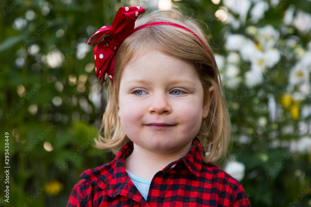 Medium horizontal portrait of pretty blue-eyed blond four-year old girl in red and black checkered shirt looking sideways with cheeky expression while standing in garden Medium horizontal portrait of pretty blue-eyed blond four-year old girl in red and black checkered shirt looking sideways with cheeky expression while standing in garden