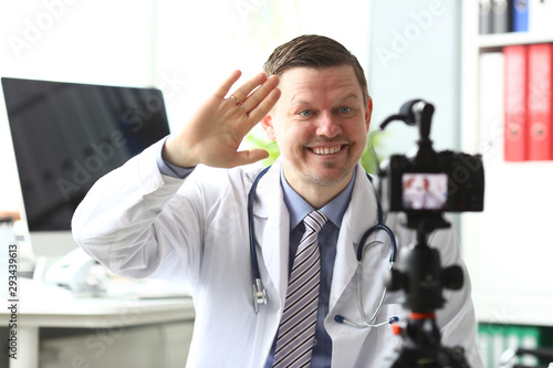 Smiling male millennial doctor wave his arm to camera saying hello to subscribers portrait