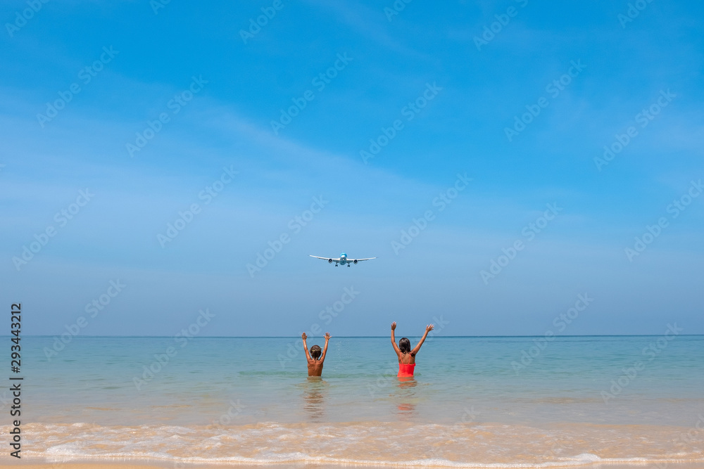 A boy and a girl have fun on the beach and greet a flying airplane with their hands. They are located on Mai Khao Beach in Thailand.