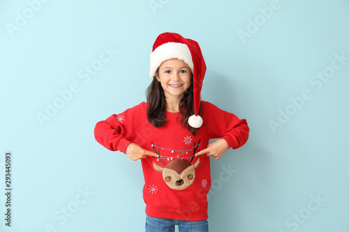 Little girl in Christmas sweater and Santa hat on color background