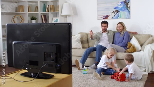 Tracking shot of happy man and woman sitting on sofa and chatting while watching TV. Adorable little girl and boy playing with toys on floor photo