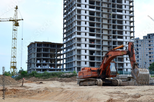 Excavator on the construction site against the background of a high multi-storey residential complex and tower cranes. Excavation, digging trenches for sewer pipes and utilities photo