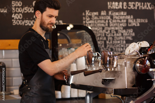 Young smiling male barista making coffee using professional coffee machine. Morning cup of coffee in caf  . Brewing coffee. Coffee shop concept.