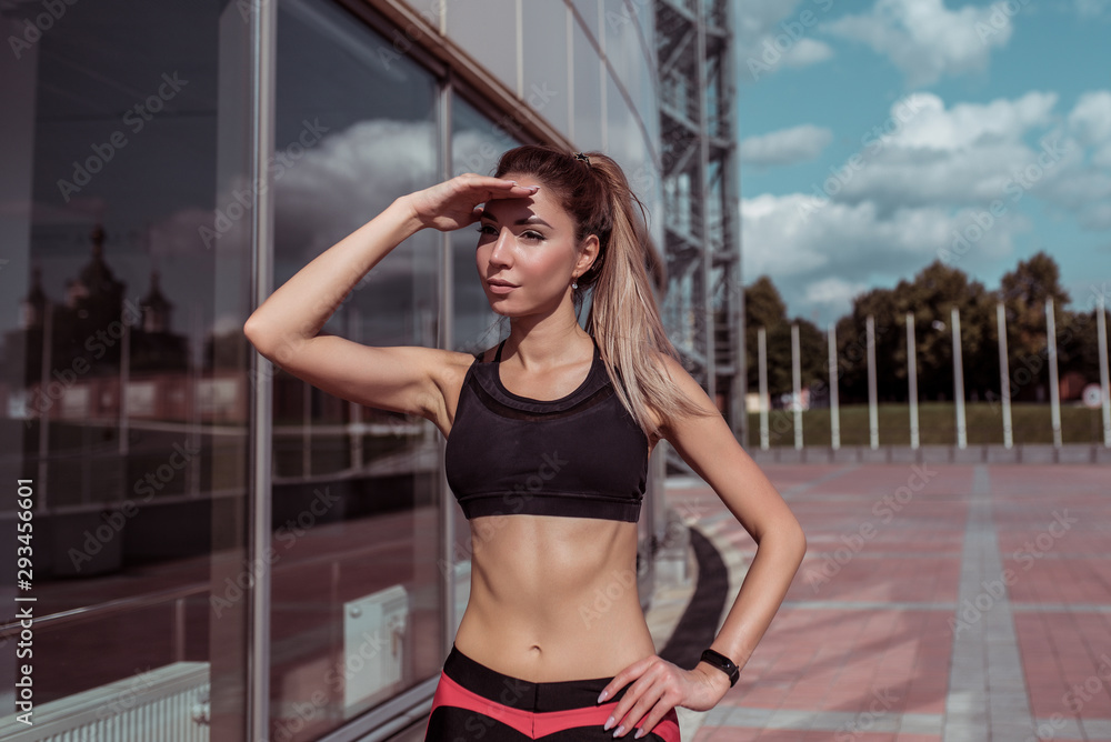 Fototapeta premium Beautiful girl athlete, covers distance into distance with hand from sun, glass windows background, summer spring city, free space motivation, sportswear, active fitness lifestyle, outdoor exercise.
