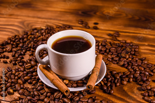 Cup of hot coffee, cinnamon sticks and scattered coffee beans on wooden table