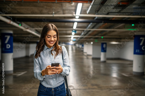Businesswoman in Underground parking with smartphone. Young woman smiling confident at underground parking lot around cars and lights. Woman in the underground car parking