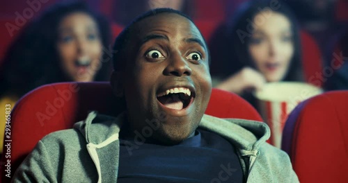Close up of the cherful and happy African American young guy sitting in the cinema while watching nice action film and making surprised and satisfied face as loves this movie and looks enthusiastic. photo