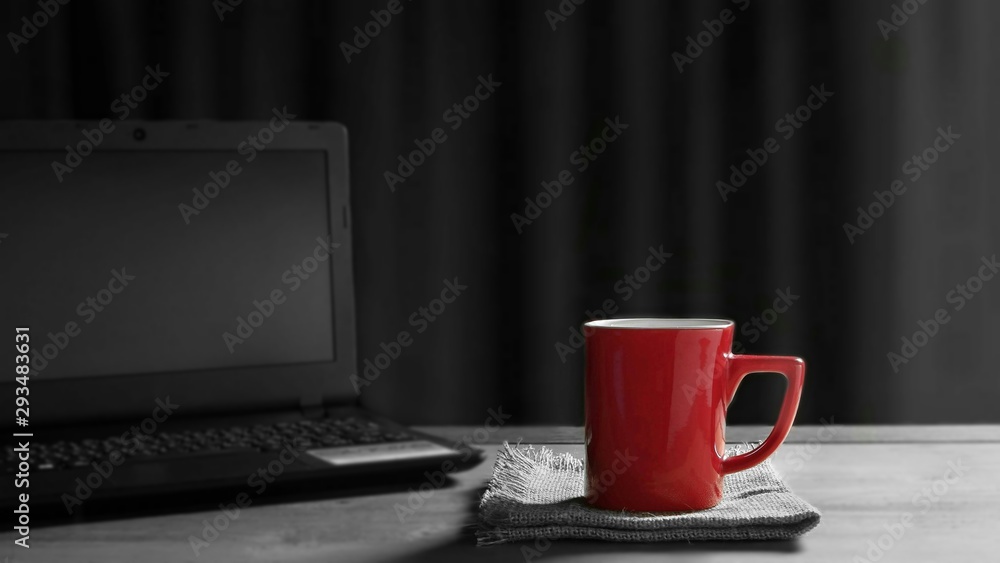 Focus on red coffee cup on napkins with blurred black laptop on wooden table and sunlight shine through the window in living room background, coffee break concept, splash red color technique Focus on red coffee cup on napkins with blurred black laptop on wooden table and sunlight shine through the window in living room background, coffee break concept, splash red color technique
