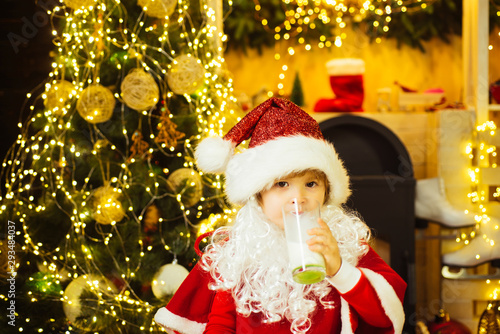 Portrait of Santa Claus Drinking milk from glass and holding cookies. Little Santa Claus kid with beard and mustache. Santa boy in Santa hat.