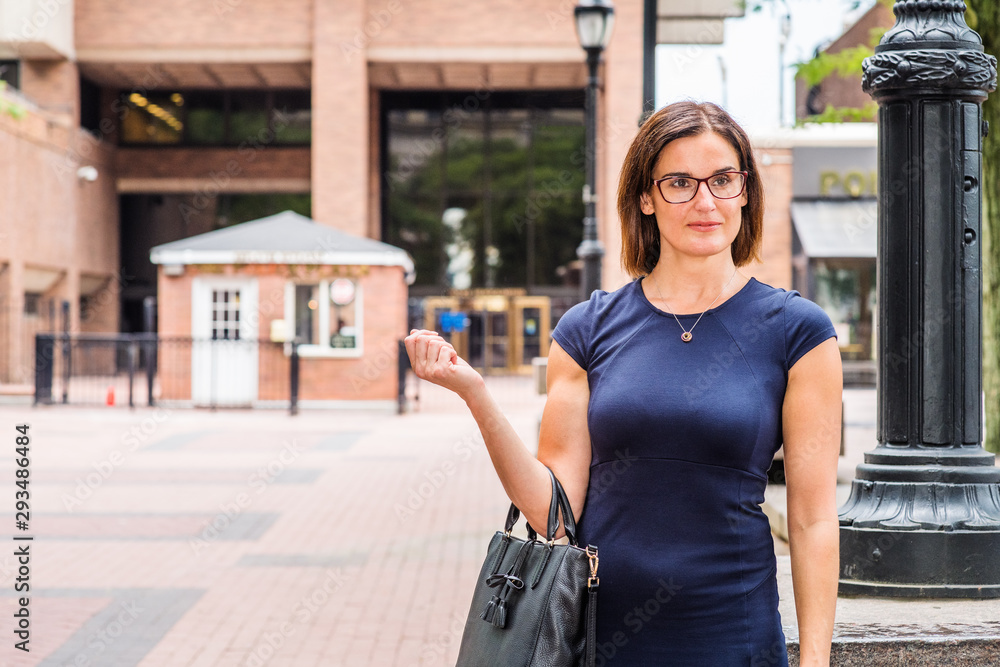 Portrait of Young Female Teacher working in New York City. Young Native American Woman wearing blue short sleeveless dress, glasses, arm carrying bag, standing on street by light pole on school campus