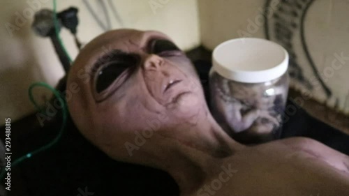 Shot panning down at the alien laying on table with creature in jar next to him and green tubes connecting to the alien. photo