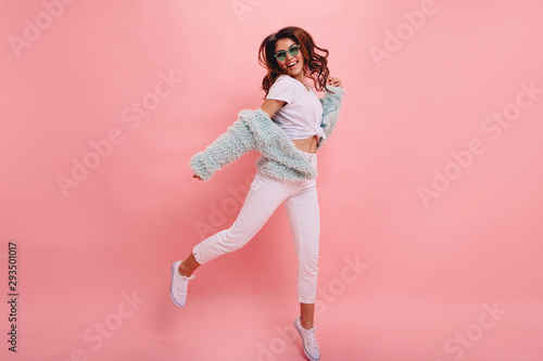  Inspired positive girl in white sneakers dancing on pink background. Gorgeous young female model with dark wavy hair jumping in studio. Not isolated. Copy space.