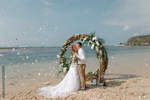 Wedding ceremony on a tropical beach in blue. Happy groom and bride kissing  under the arch decorated with flowers on the sandy beach. Rose petals fall from above. Wedding and honeymoon concept.
