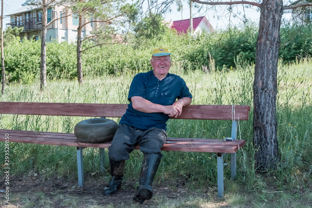 An elderly man sits on a wooden bench in rubber boots. Old man in the summer garden An elderly man sits on a wooden bench in rubber boots. Old man in the summer garden