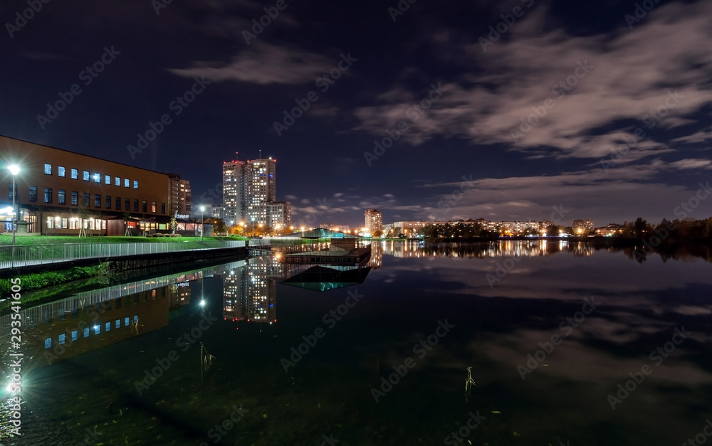 Night city landscape - high-rise houses with lighted windows on the shore of the pond, dark sky with clouds and reflection in the water