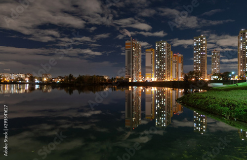 Night city landscape - high-rise houses with lighted windows on the shore of the pond, dark sky with clouds and reflection in the water
