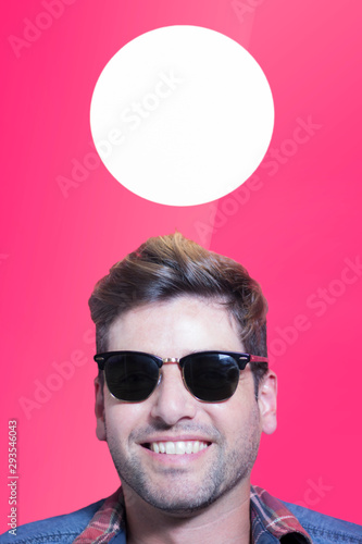 Close up portrait of a smiling young man wearing sunglasses over pink background and a round light above his head. Illuminated blank lamp with copy space. Mock up.