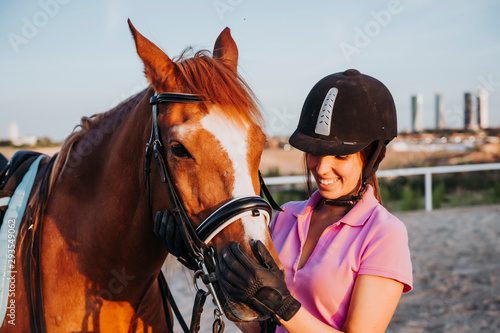 Young smiling woman standing in relax outside with wonderful chestnut horse on bridle and looking away. © karrastock