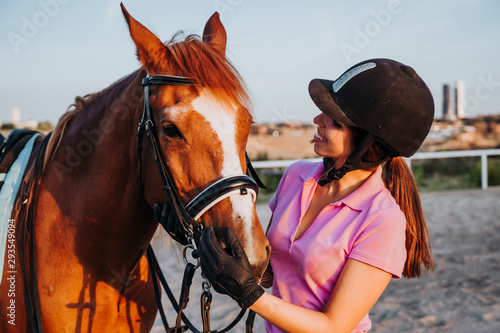 Young smiling woman standing in relax outside with wonderful chestnut horse on bridle and looking away. © karrastock