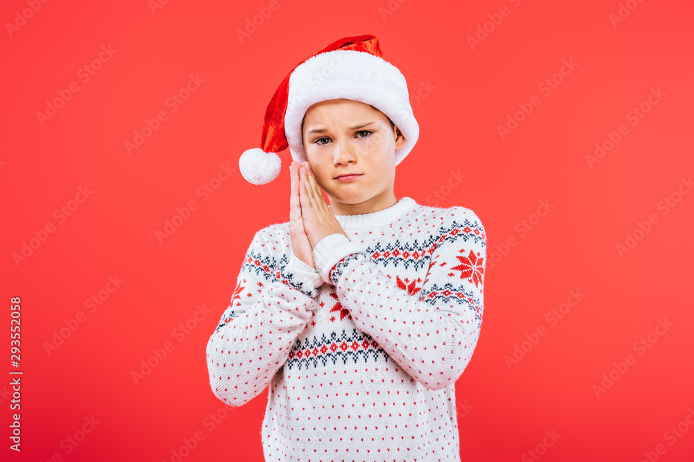 sad kid in sweater and santa hat showing please gesture isolated on red sad kid in sweater and santa hat showing please gesture isolated on red