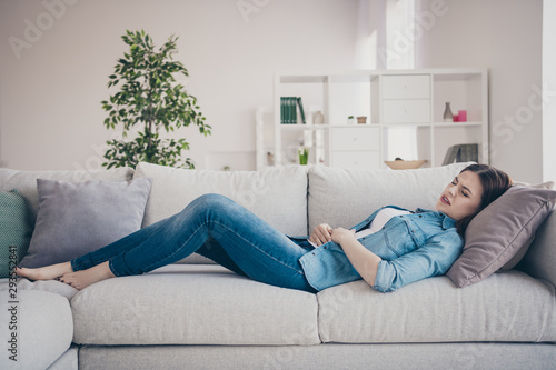 Profile photo of pretty lady suffering from pms holding hands on hurted belly lying sofa trying to relax wearing jeans outfit apartment indoors