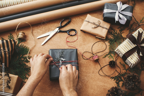Wrapping christmas gifts flat lay. Hands wrapping stylish christmas gift box in black paper and scissors, rustic presents, thread, pine branches and cones on wooden table photo