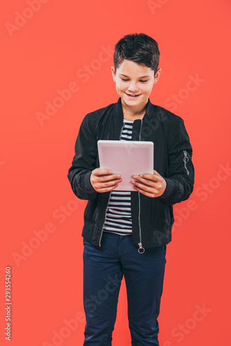 smiling kid in jacket using digital tablet isolated on red