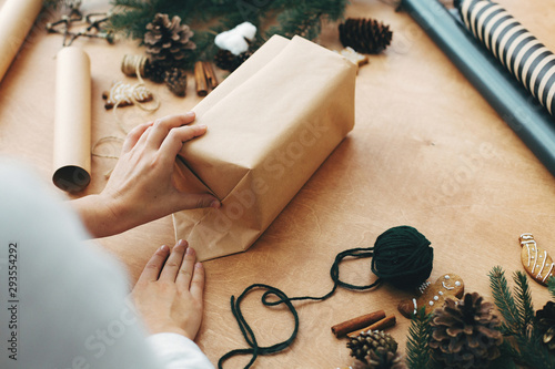 Hands wrapping christmas gift in craft paper and pine branches, cones, gingerbread cookies, thread, cinnamon on rural wooden table. Wrapping stylish christmas gifts concept photo