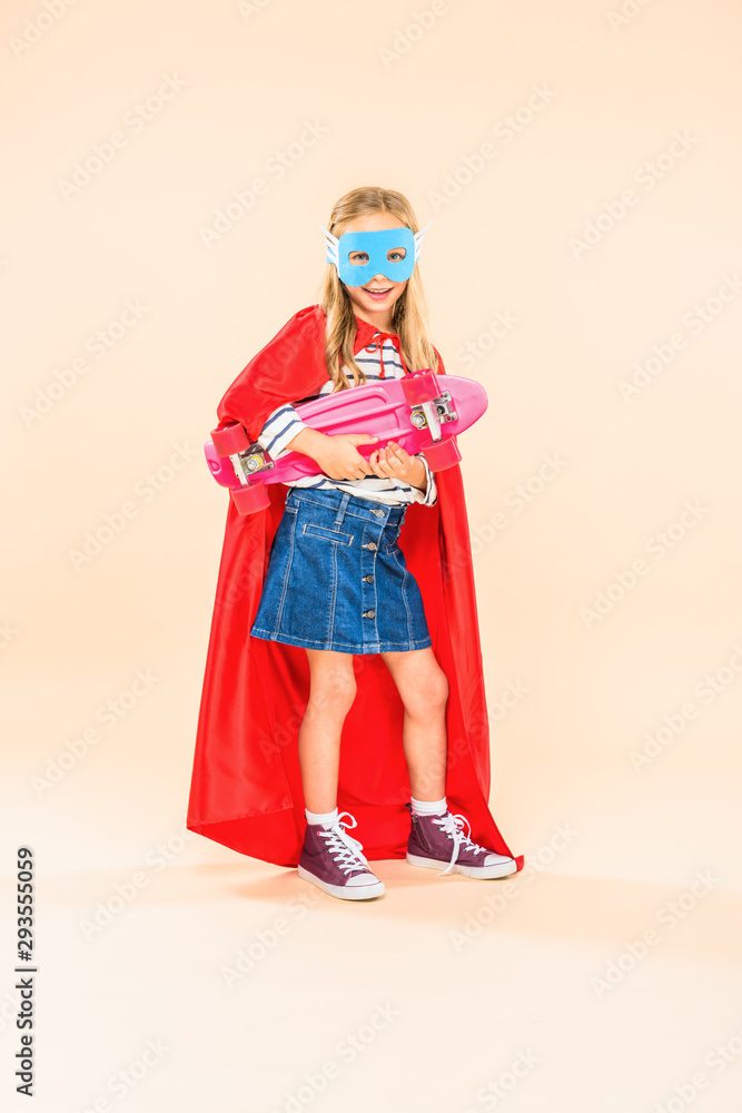 Full length view of child in mask and hero cloak holding skateboard on pink
