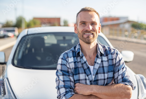 An attractive young man stands near a white car.
