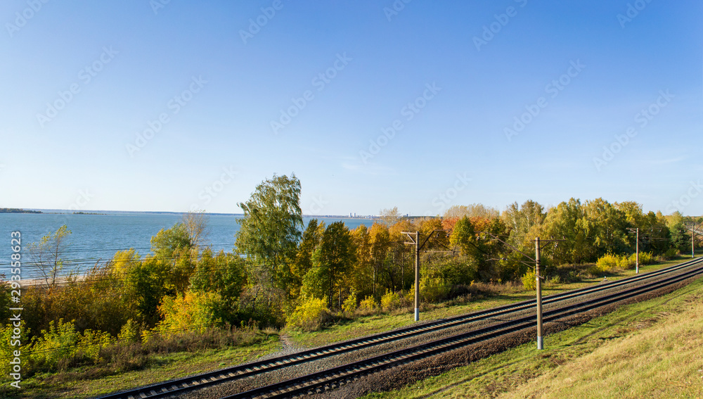 Railway on the background of the river Bank on an autumn Sunny day. Beautiful yellow-blue panorama of trees by the water. Railway on the background of the river Bank on an autumn Sunny day. Beautiful yellow-blue panorama of trees by the water.