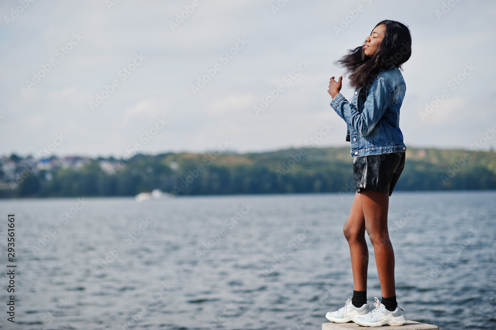 African american dark skinned slim model posed in a black shorts and jeans jacket against sea side. African american dark skinned slim model posed in a black shorts and jeans jacket against sea side.